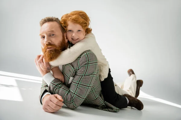 Red haired boy in beige jumper hugging stylish father while lying on grey background — Stock Photo