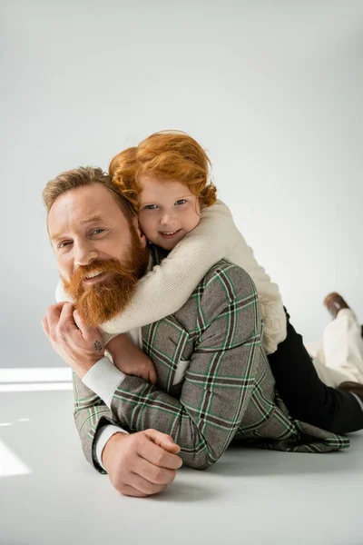 Cheerful redhead boy embracing bearded father lying on grey background — Stock Photo