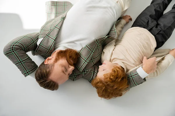 Top view of redhead father and son in trendy outfit looking at each other while lying on grey background — Stock Photo