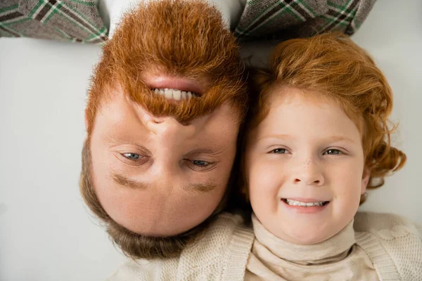 Vista superior do menino ruivo com o pai barbudo sorrindo para a câmera enquanto deitado no fundo cinza — Fotografia de Stock