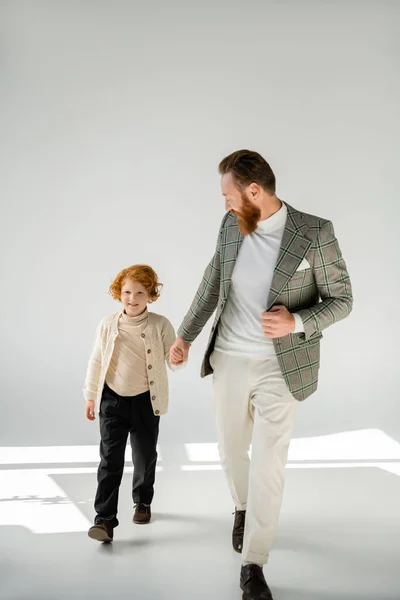 Full length of trendy bearded man holding hands with redhead son and walking on grey background — Stock Photo