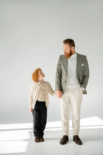 Bärtiger Mann im karierten Blazer mit rothaarigem Sohn in Strickjacke und Händchen haltend auf grauem Hintergrund — Stockfoto