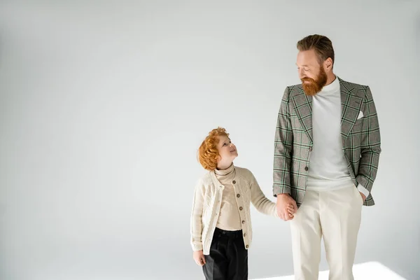 Рудий батько і син в модному вбранні тримаються за руки і дивляться один на одного на сірому фоні — стокове фото