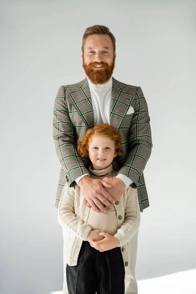 Felice uomo barbuto in plaid blazer abbracciando il figlio rossa e sorridendo alla fotocamera su sfondo grigio — Foto stock