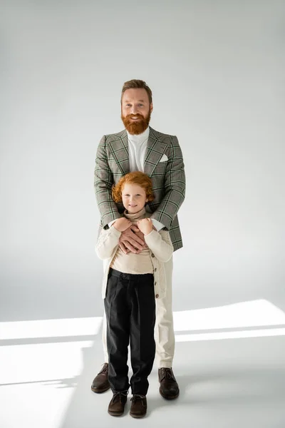 Повна довжина рудого хлопчика, що стоїть біля бородатого батька, обіймає його на сірому фоні — стокове фото