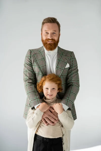 Happy and stylish dad and son with red hair smiling at camera on grey background — Stock Photo