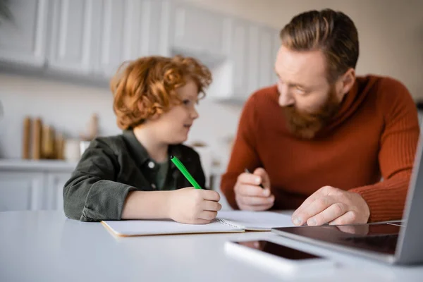 Бородатий чоловік розмовляє з рудим сином робить домашнє завдання біля розмитого ноутбука — стокове фото