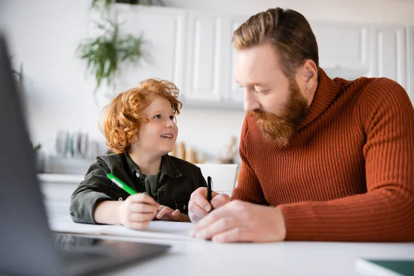 Happy boy looking at bearded father while doing homework near blurred laptop — Stock Photo