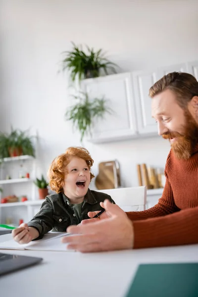 Бородатый мужчина разговаривает со смеющимся рыжим сыном, который делает домашнее задание на кухне — стоковое фото