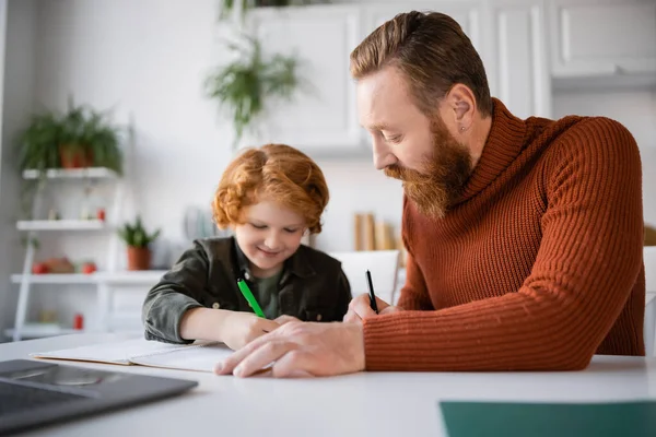 Smiling kid writing in notebook near bearded dad and laptop on blurred foreground — Stock Photo