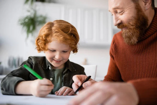 Lächelndes rothaariges Kind, das in Notizbuch schreibt, während es in der Nähe seines Vaters Hausaufgaben macht — Stockfoto