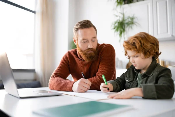 Redhead child writing in notebook near bearded dad and laptop — Stock Photo