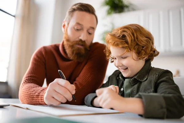 Uomo barbuto che tiene penna vicino notebook e sorridente rosso figlio facendo i compiti — Foto stock