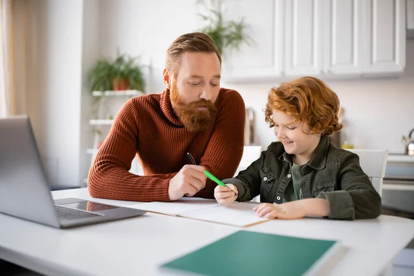 Bärtiger Mann hilft lächelndem rothaarigem Sohn beim Schreiben in Notizbuch neben verschwommenem Laptop — Stockfoto