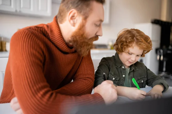 Bearded man looking at redhead son writing in notebook while doing homework on blurred foreground — Stock Photo