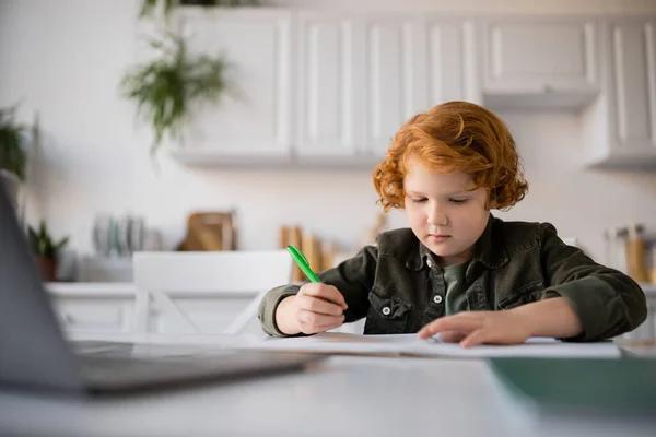 Redhead child holding pen while learning at home near blurred laptop — Stock Photo