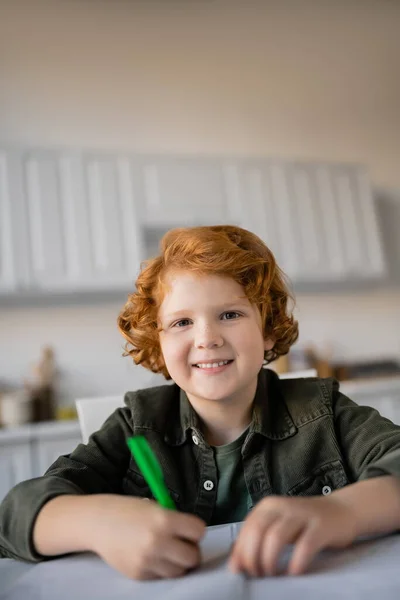 Heureux rousse enfant avec stylo regardant caméra tout en faisant des devoirs sur le premier plan flou — Photo de stock