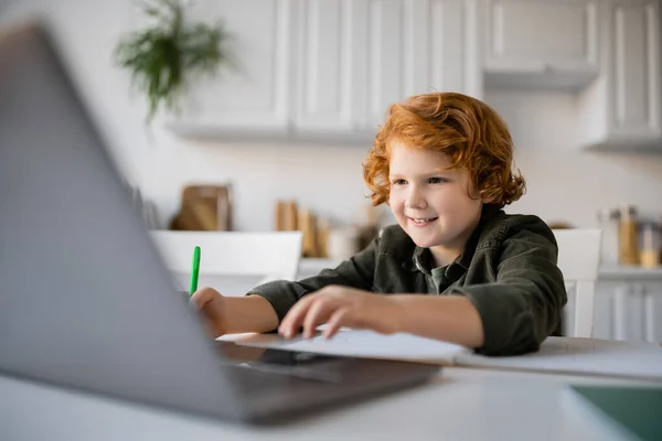 Веселый рыжий мальчик делает домашнее задание рядом с размытым ноутбуком на кухне — стоковое фото