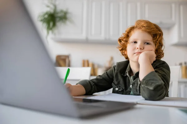 Annoiato rossa ragazzo seduto con mano vicino al viso mentre fa i compiti a casa al computer portatile offuscato — Foto stock