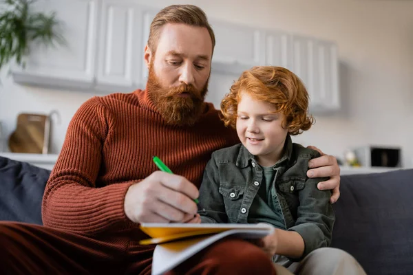 Bärtiger Mann schreibt in Notizbuch, während er auf Sofa neben lächelndem rothaarigem Sohn sitzt — Stockfoto