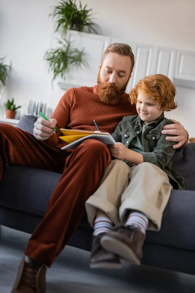 Рыжий мальчик пишет в блокноте возле бородатого папы на диване дома — стоковое фото