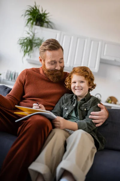 Щасливий бородатий чоловік обіймає веселий рудий син сидить на дивані з блокнотом — стокове фото