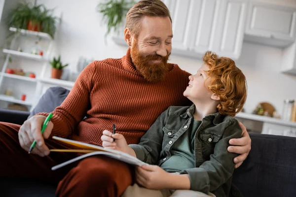 Рыжий отец и сын сидят на диване с ручками и блокнотом и улыбаются друг другу. — стоковое фото