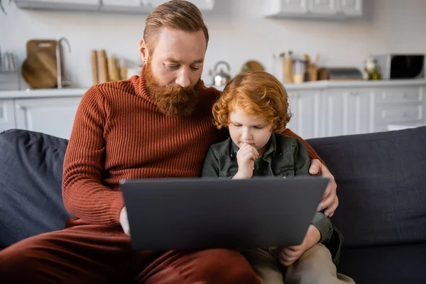 Nachdenkliches rothaariges Kind blickt auf Laptop, während es neben bärtigem Papa auf der Couch sitzt — Stockfoto
