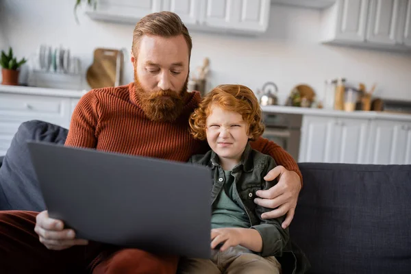 Bored boy frowning near laptop and redhead dad on couch at home — Stock Photo