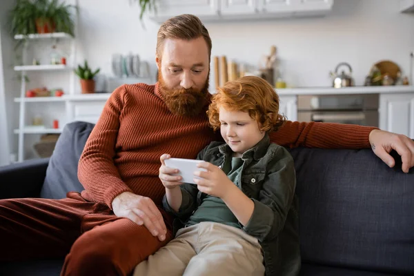 Рыжий мальчик сидит на диване рядом с бородатым отцом и использует смартфон — стоковое фото