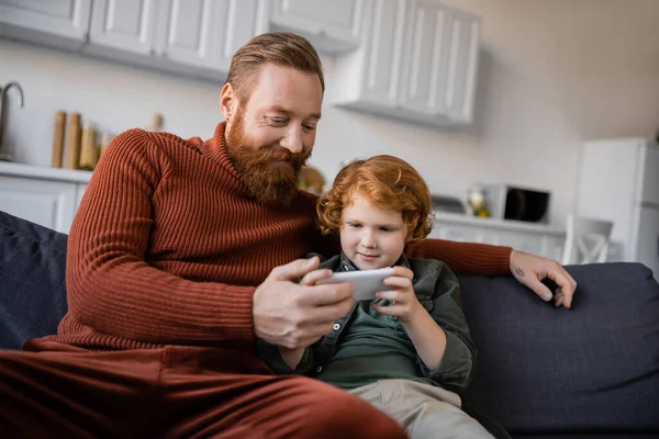 Улыбающийся бородатый мужчина сидит на диване возле рыжеволосого сына с помощью мобильного телефона — стоковое фото