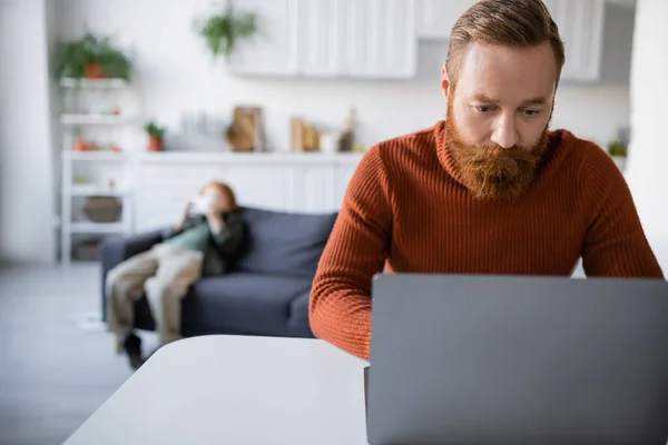 Rossa e barbuto uomo lavorando su laptop vicino al figlio su sfondo sfocato — Foto stock