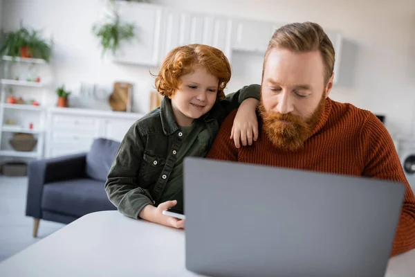 Рыжий ребенок улыбается рядом с бородатым отцом, работающим над размытым ноутбуком дома. — стоковое фото