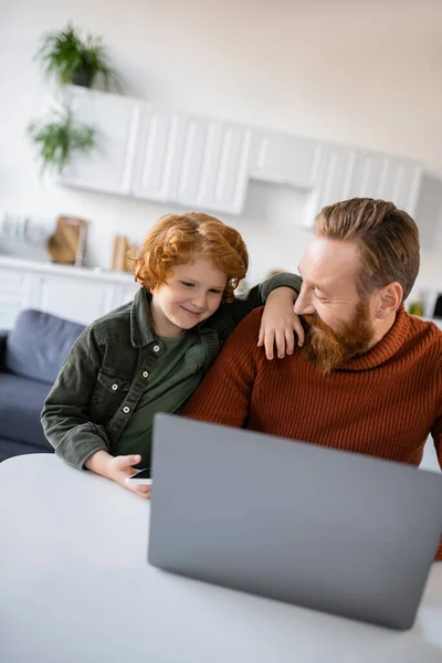 Happy boy standing near smiling father working on blurred laptop at home — Stock Photo