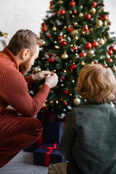 Vista trasera del papá pelirrojo y el hijo decorando borrosa árbol de Navidad en casa - foto de stock