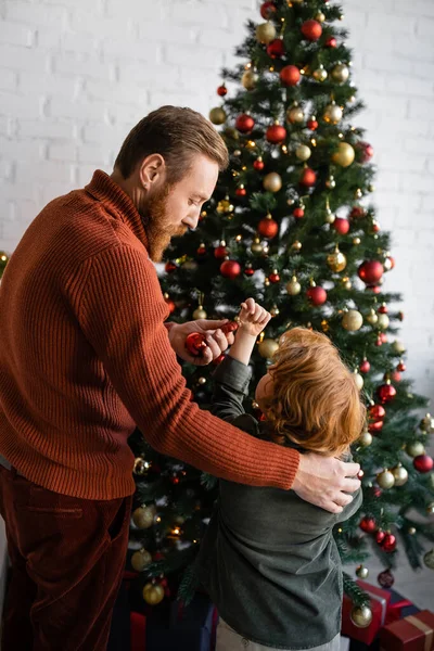 Bärtiger Mann hält Christbaumkugel neben rothaarigem Sohn und schmückt Weihnachtsbaum — Stockfoto