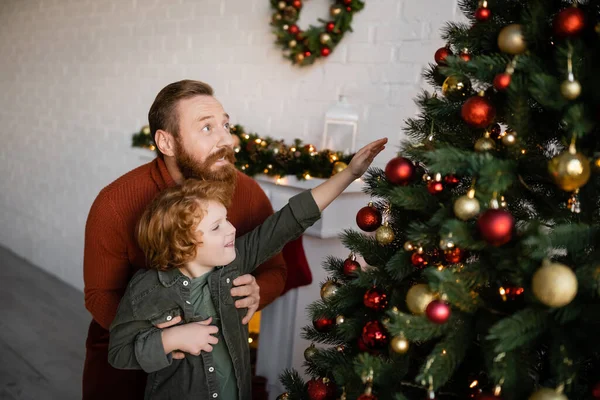Bearded man holding son reaching baubles on decorated Christmas tree — Stock Photo