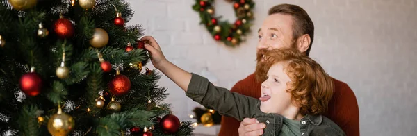 Ruiva menino saindo língua e decoração árvore de Natal perto pai barbudo, bandeira — Fotografia de Stock