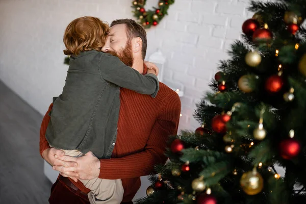 Ruiva menino abraçando pai segurando-o perto decorado árvore de Natal — Fotografia de Stock