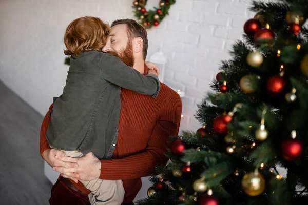 Redhead boy hugging dad holding him near decorated Christmas tree — Stock Photo
