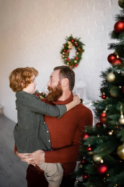 Cheerful bearded man holding redhead son in hands near blurred pine tree decorated with Christmas baubles — Stock Photo