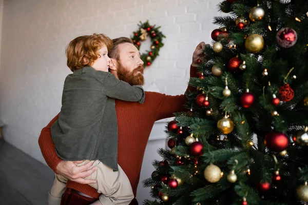 Bärtiger Mann hält rothaarigen Sohn in den Händen, während er zu Hause den Weihnachtsbaum schmückt — Stockfoto