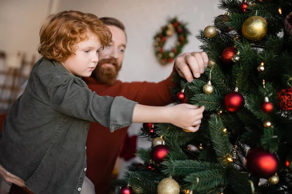 Verschwommener Mann hält rothaarigen Sohn beim Dekorieren von Fichten mit Weihnachtskugeln zu Hause — Stockfoto