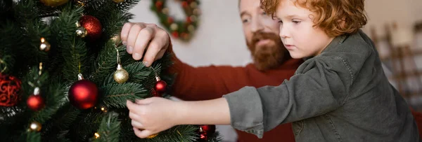 Bambino rosso che decora l'albero di Natale vicino al padre barbuto sfocato, striscione — Foto stock