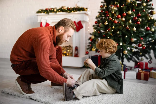 Vista lateral del niño pelirrojo sentado en el suelo cerca de papá atando sus cordones y el árbol de Navidad con chimenea decorada - foto de stock