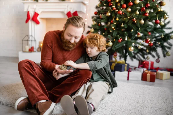 Рыжий отец и ребенок держат рождественскую безделушку на полу в гостиной с праздничным декором — стоковое фото