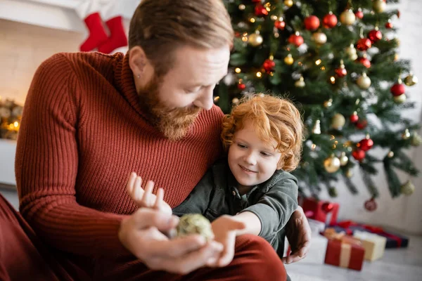 Barbudo hombre sosteniendo borrosa chuchería cerca pelirroja hijo y árbol de Navidad en el fondo - foto de stock