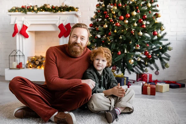 Rothaarige Vater und Sohn sitzen mit gekreuzten Beinen auf dem Boden im Wohnzimmer mit Weihnachtsbaum und dekoriertem Kamin — Stockfoto