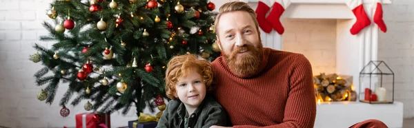 Criança e pai com cabelo vermelho sorrindo para a câmera perto da árvore de Natal e lareira, banner — Fotografia de Stock