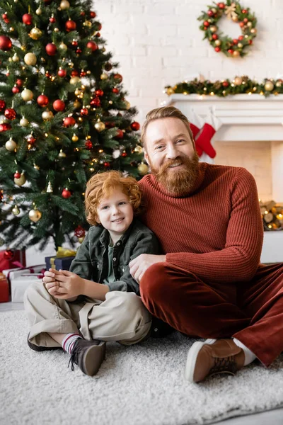 Веселый отец и ребенок с рыжими волосами сидят на полу возле елки и смотрят в камеру — стоковое фото